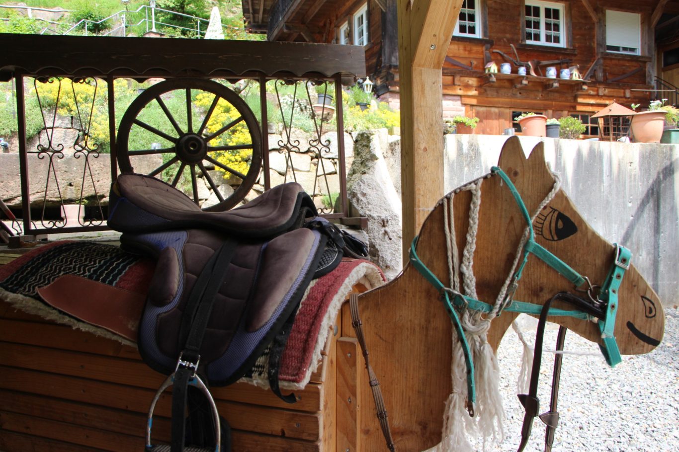 Sattel auf einem hölzernen Pferd, im Hintergrund Garten und Wagenrad sichtbar.