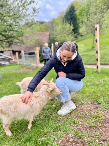 Junge Frau streichelt eine Ziege auf einer grünen Wiese. Im Hintergrund sind weitere Personen.