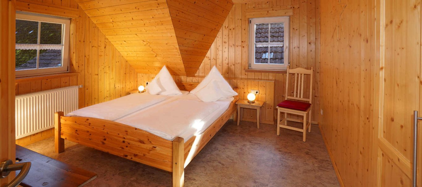 Ferienwohnung Blumenwiese Helles Holzzimmer mit Doppelbett, zwei Kissen und einem Stuhl.